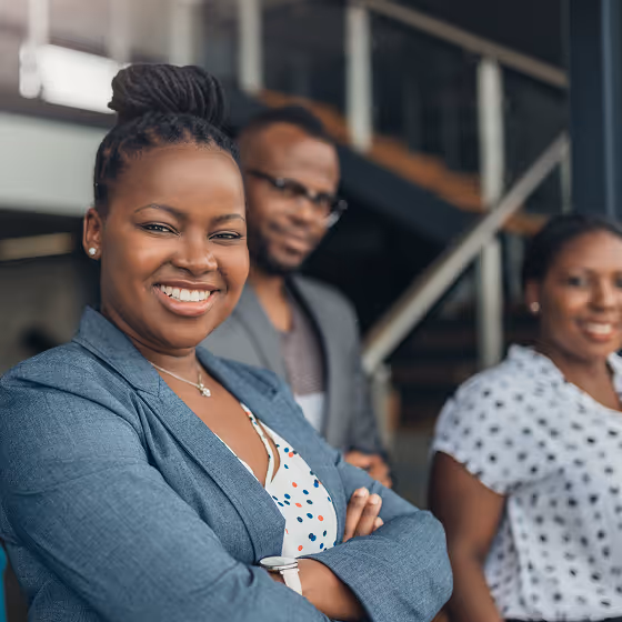 Smiling woman with braided hair and a blue blazer standing confidently with folded arms, accompanied by two smiling colleagues in the background.