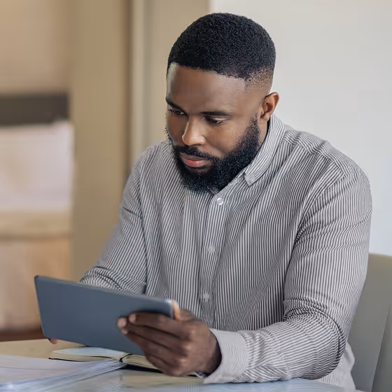Man with a beard wearing a striped shirt sitting at a desk and looking at a tablet.